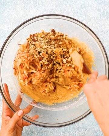 two hands mixing apple muffin batter in a large glass bowl.