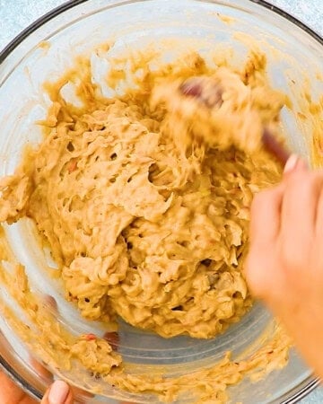 two hands mixing apple muffin batter in a large glass bowl.
