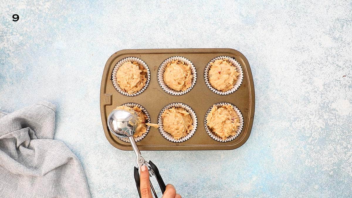 a hand adding batter into a metal muffin pan using an ice cream scoop.