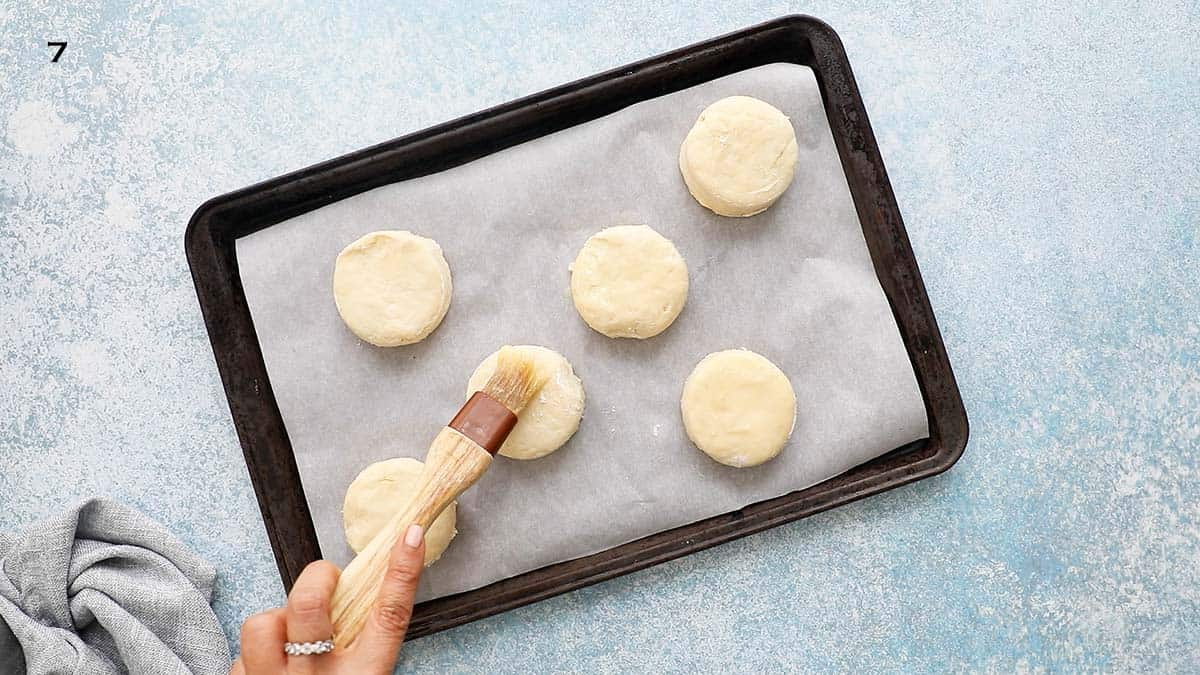 a hand brushing melted butter on cut biscuit dough placed on a baking sheet.