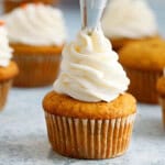a hand piping white frosting on top of a pumpkin cupcake.
