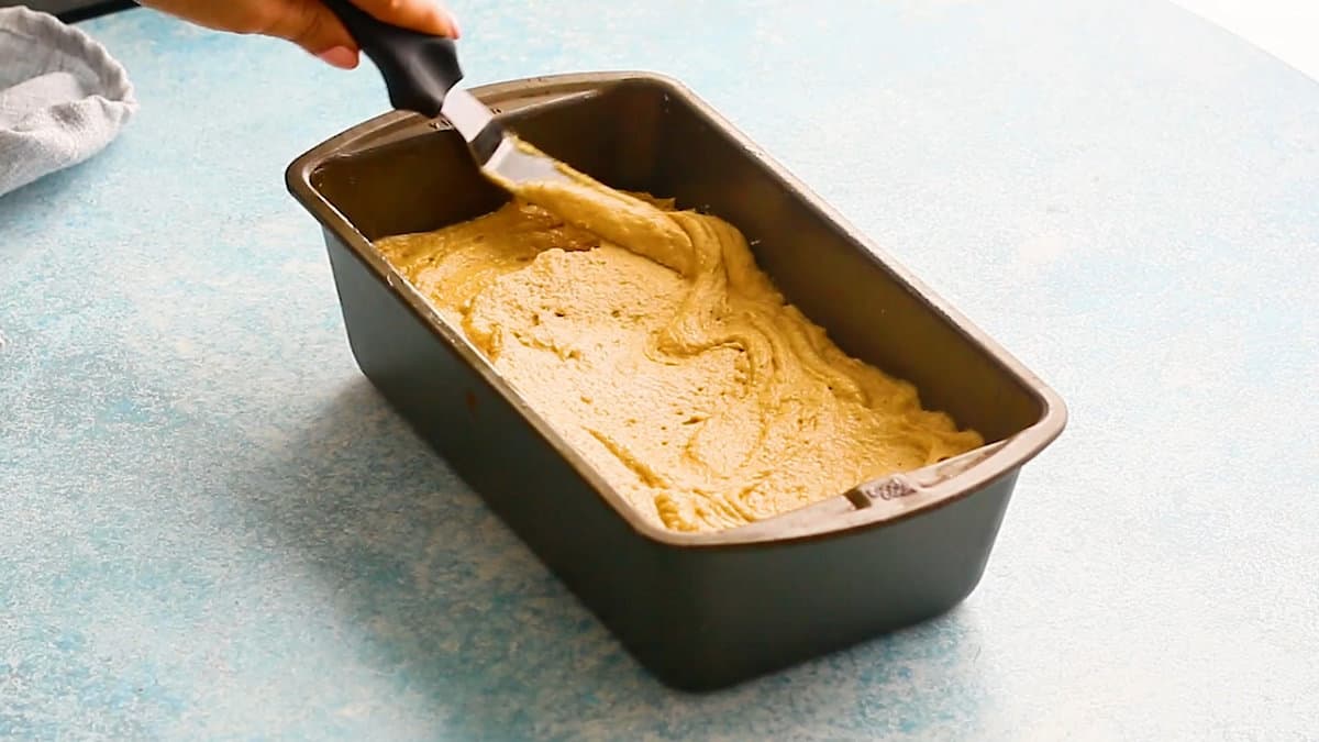 a hand spreading pumpkin cake batter in a loaf an with a flat spatula.