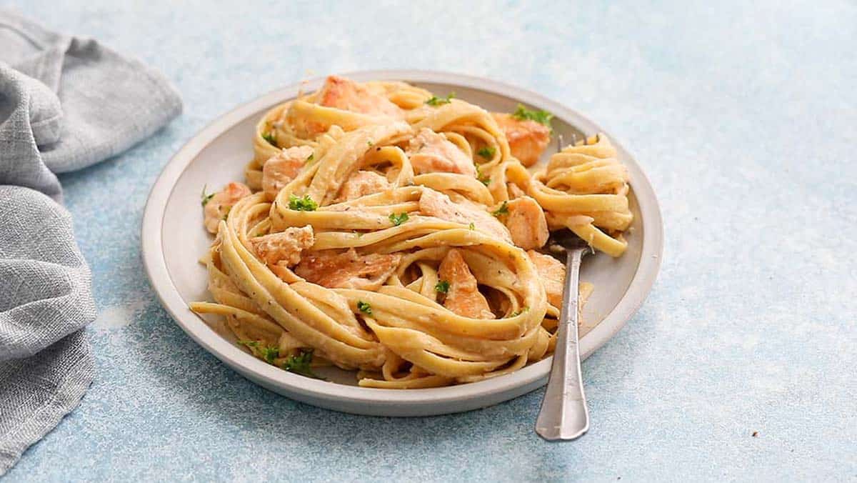 a white plate with cooked fettuccini and salmon along with a fork.