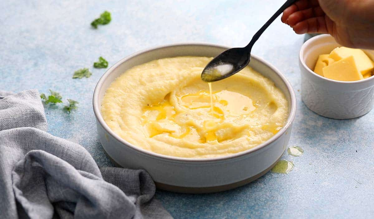 a hand drizzling melted butter using a black spoon over mashed potatoes in a white bowl.
