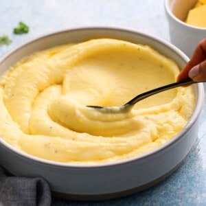 a hand swirling mashed potatoes in a white bowl using a black spoon.