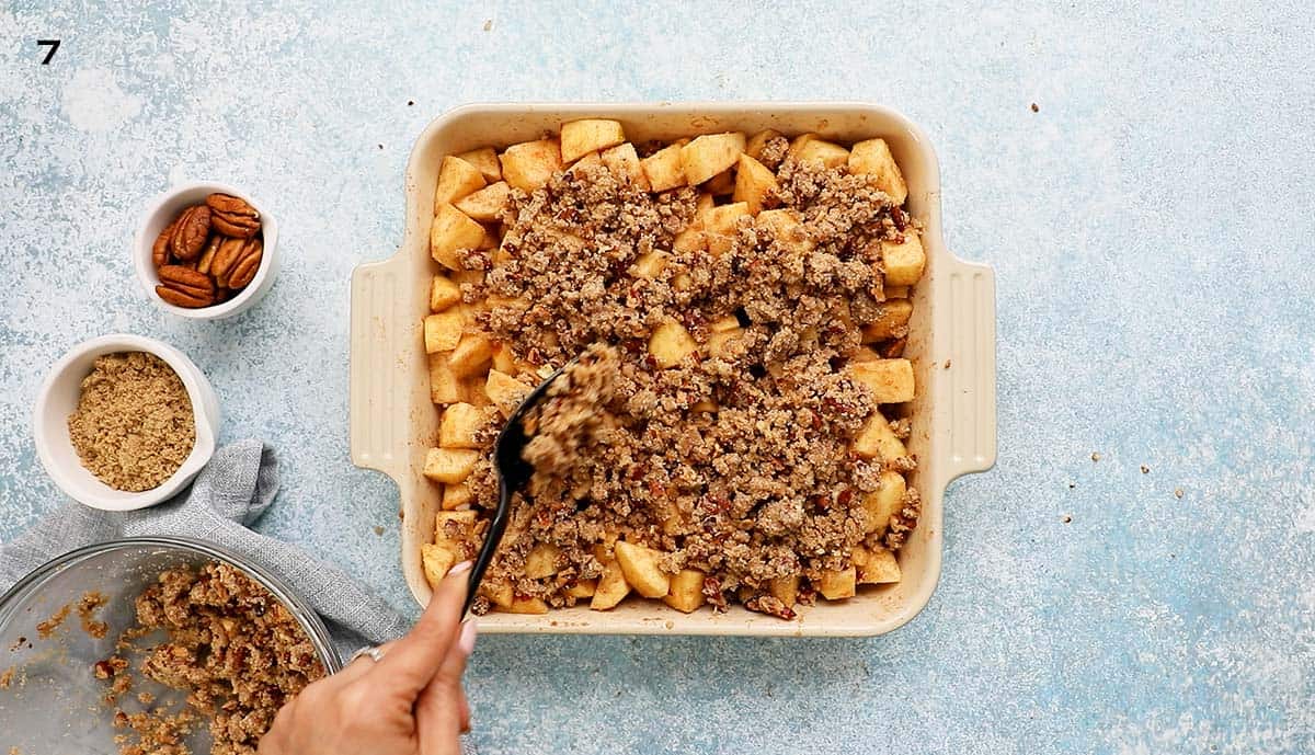 a hand sprinkling crumble topping on top of chopped apples in a square baking dish.