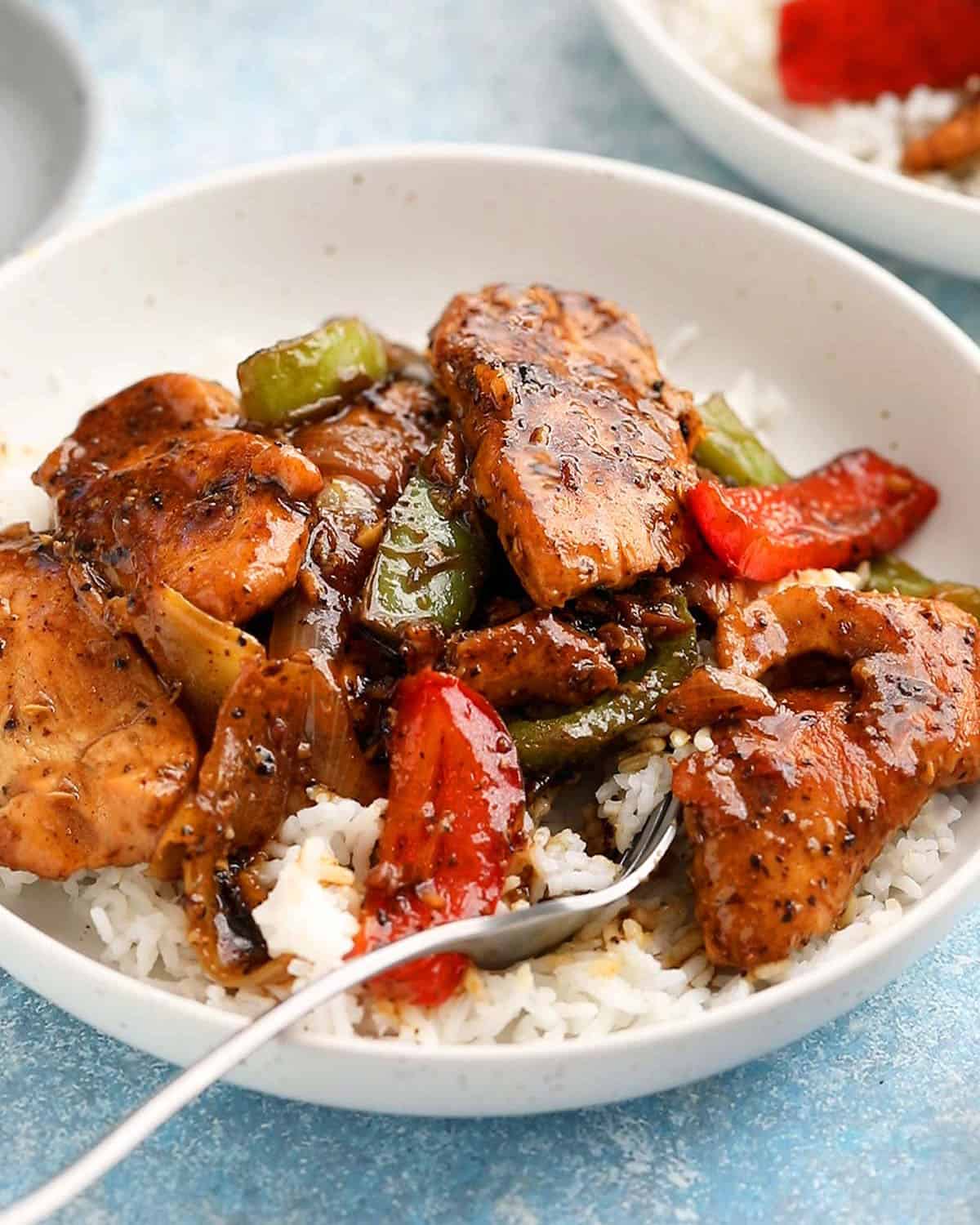 black pepper chicken stir fry with white rice in a white bowl and a metal fork.