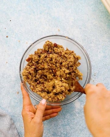 two hands mixing apple crumble topping in a small glass bowl.