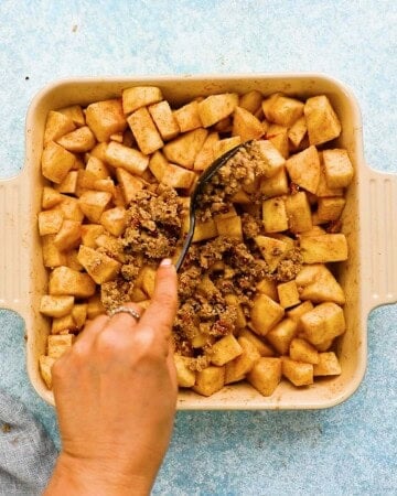 a hand adding apple crumble topping into a square baking dish.