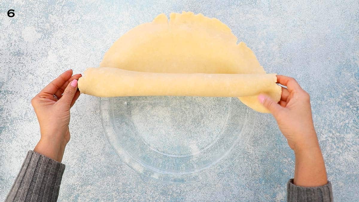 two hands placing a rolled pie crust over a glass pie plate. 