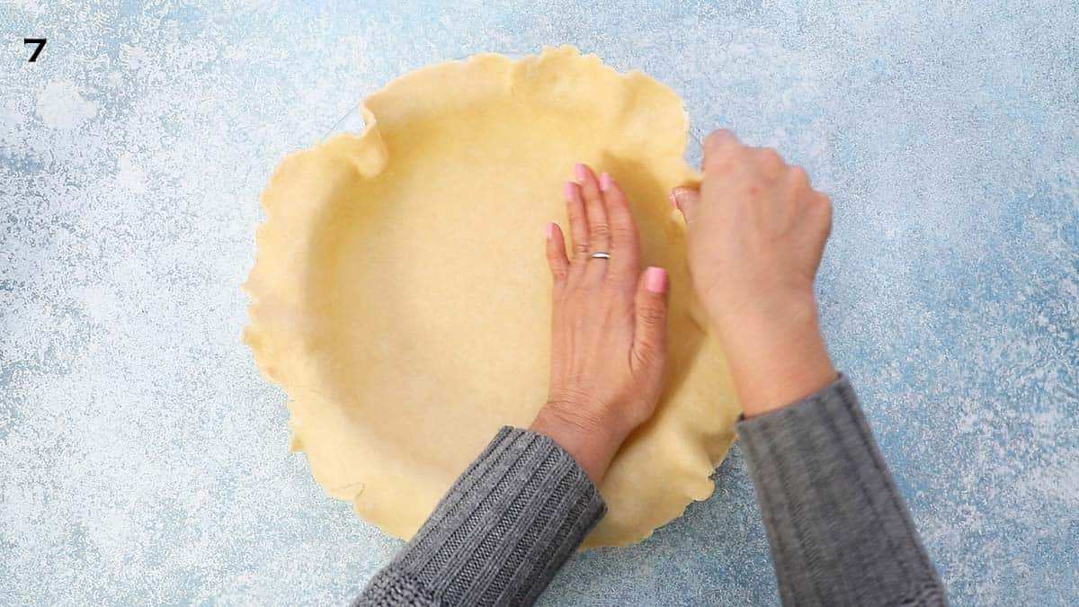 two hands shaping a rolled pie crust into a glass pie plate.