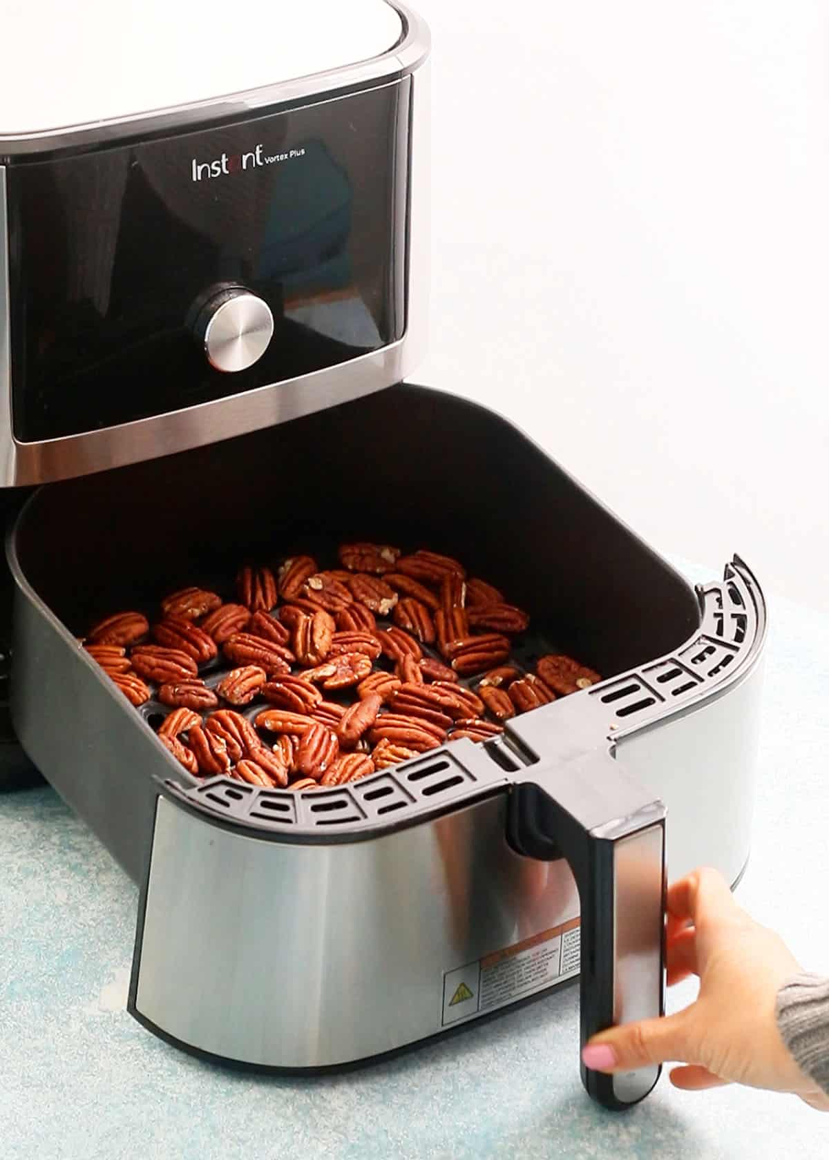 a hand holding the handle of an air fryer basket filled with toasted pecans.