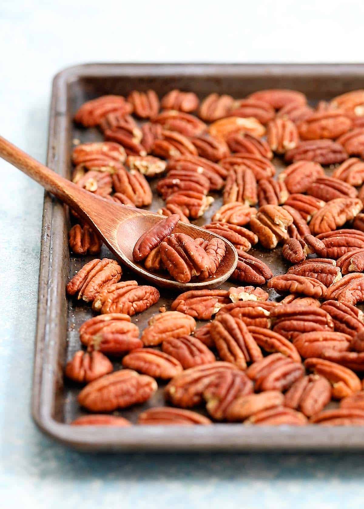 a black baking sheet filled with toasted pecans along with a wooden spoon.