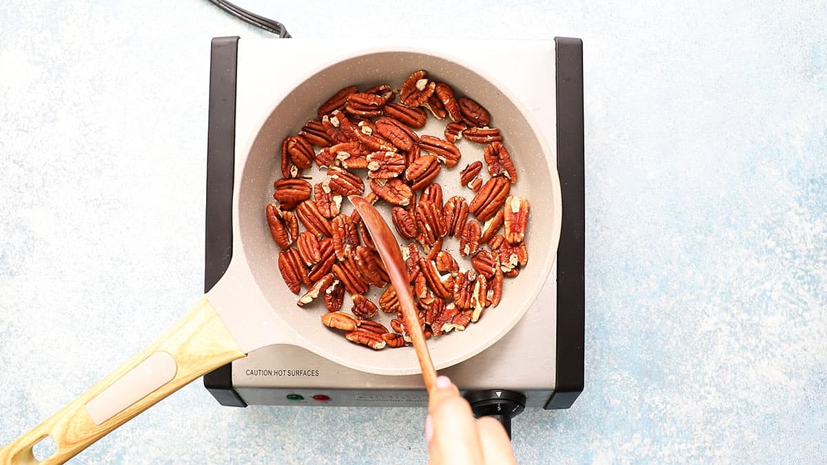 a hand stirring pecans in a small white skillet using a wooden spoon.