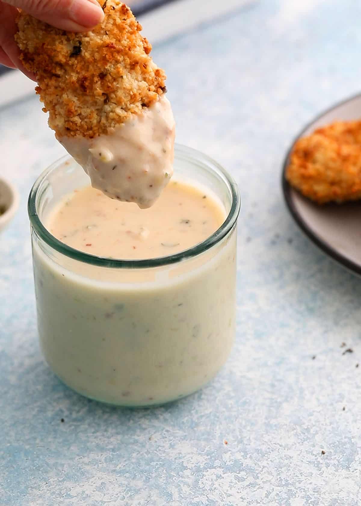 a hand holding one breaded chicken tender dipped in white sauce.