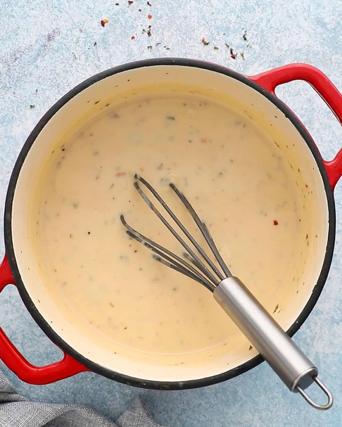 a white pot with white sauce along with a black whisk.