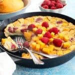 yellow mango clafoutis with pink raspberries in a black skillet.
