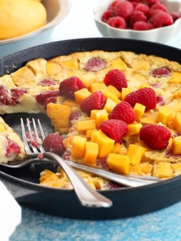 yellow mango clafoutis with pink raspberries in a black skillet.