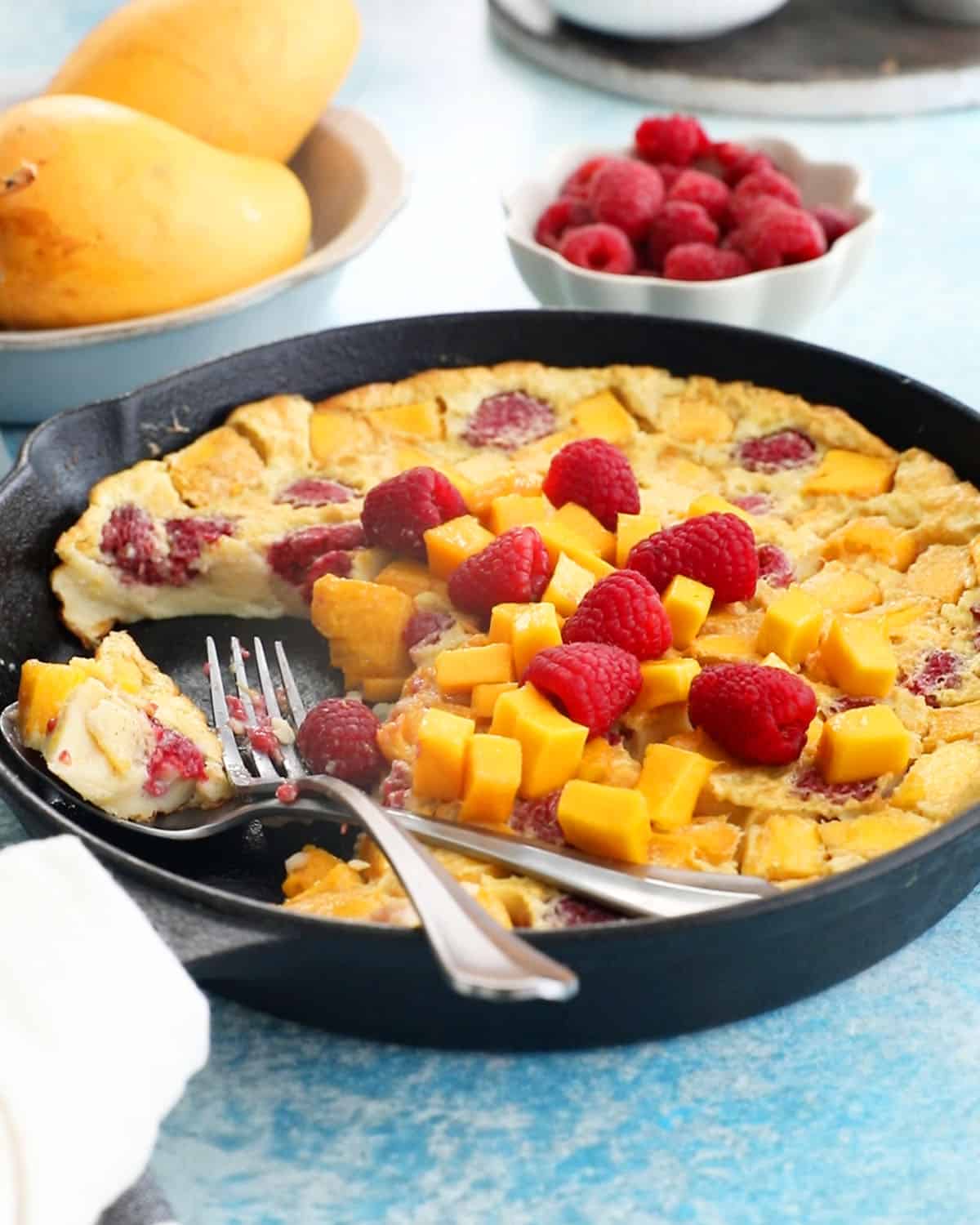yellow mango clafoutis with pink raspberries in a black skillet.