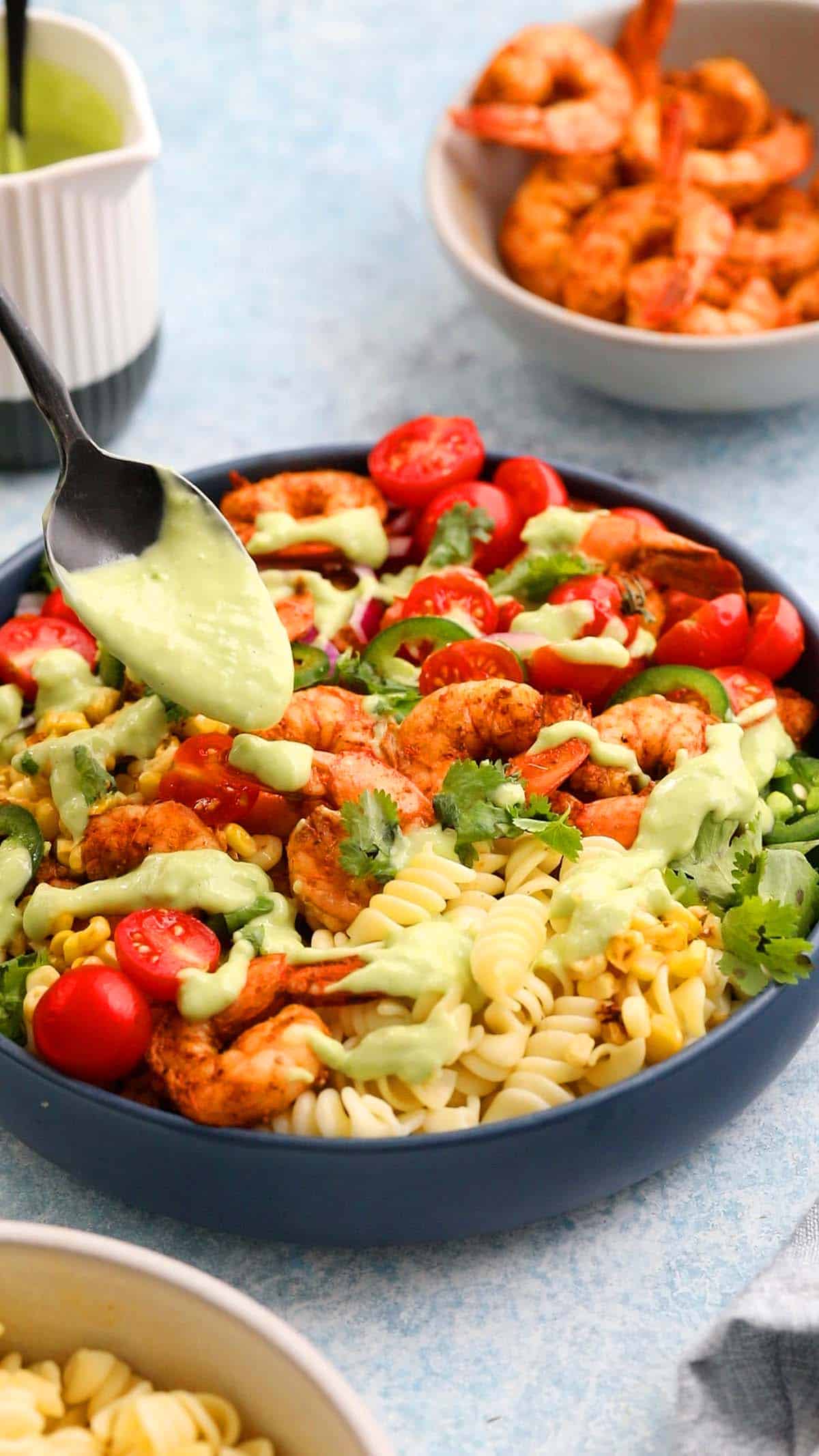 a hand drizzling green sauce on top of assembled shrimp and pasta salad in a blue bowl.