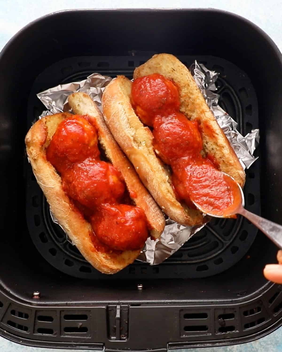 a hand spreading red sauce over chicken meatball subs in a black air fryer basket.
