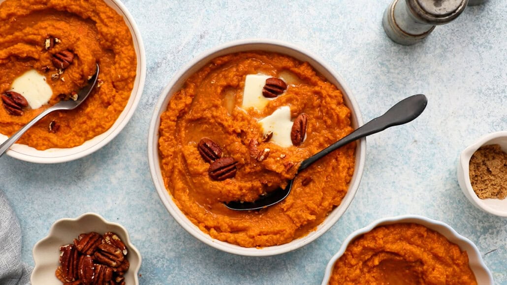 A bowl of sweet potato puree topped with melted butter and pecans sits on a light blue surface. A spoon rests in the bowl. Surrounding it, there are smaller bowls filled with more puree, pecans, and brown sugar. A pepper grinder is also visible nearby.