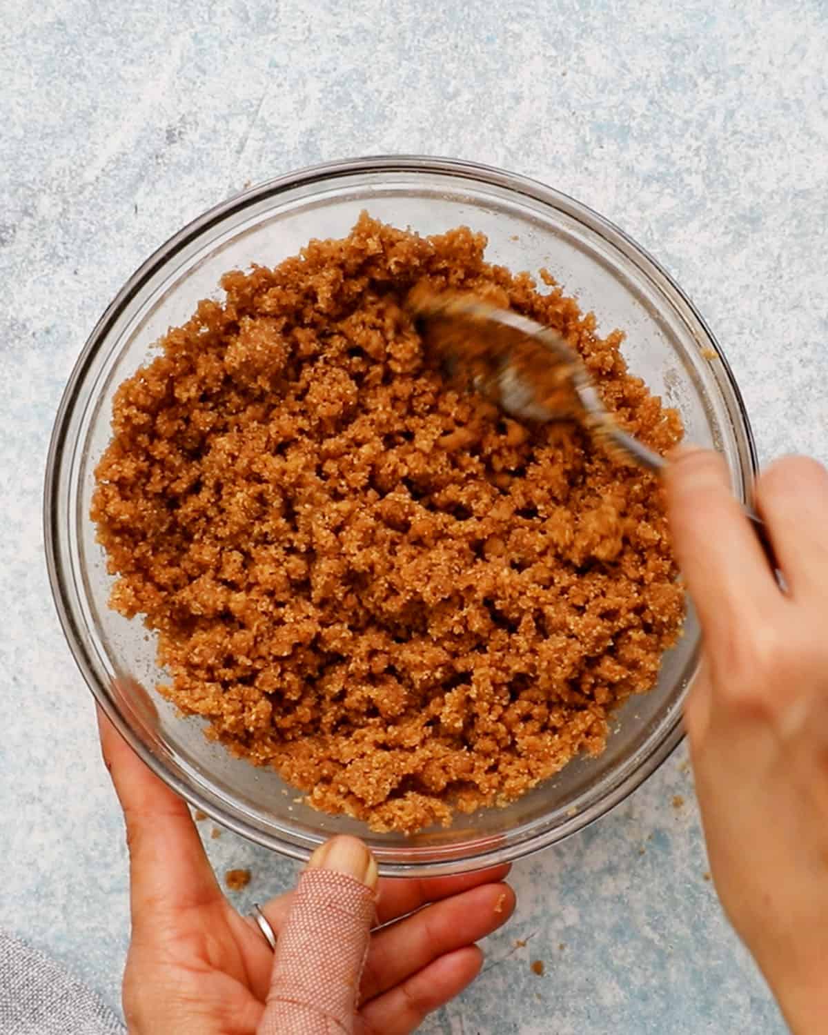 two hands mixing brown cracker crumbs in a glass bowl.