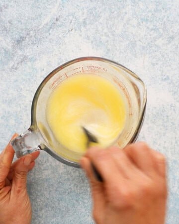 two hands mixing yellow liquid in a glass measuring cup.