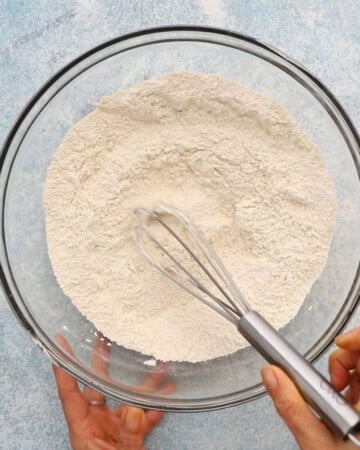 two hands whisking white flour in a large glass bowl.