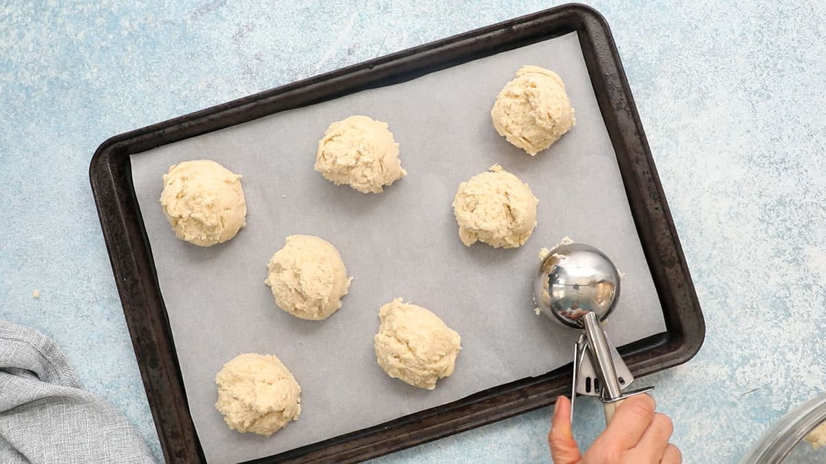 a hand placing a scoop of biscuit dough in a parchment lined baking sheet.
