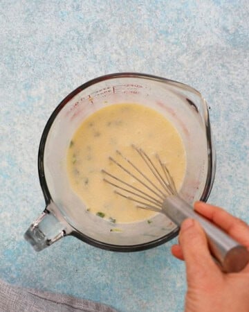 two hands whisking yellow liquid in a glass measuring cup.