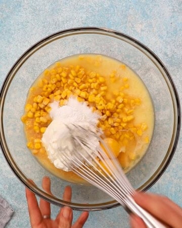 two hands whisking corn casserole mixture in a large glass bowl.