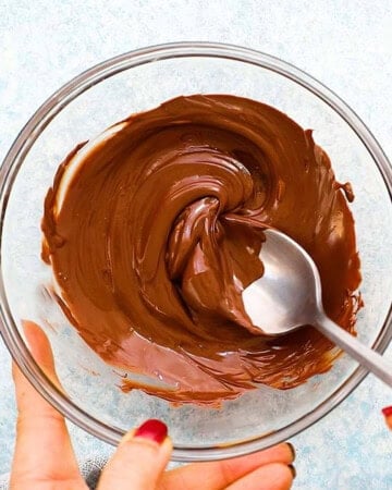 two hands mixing melted brown chocolate in a glass bowl using a spoon.