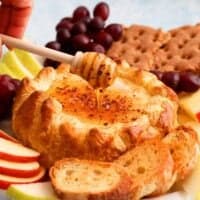 a hand holding one wooden honey dipper above a round baked brie.