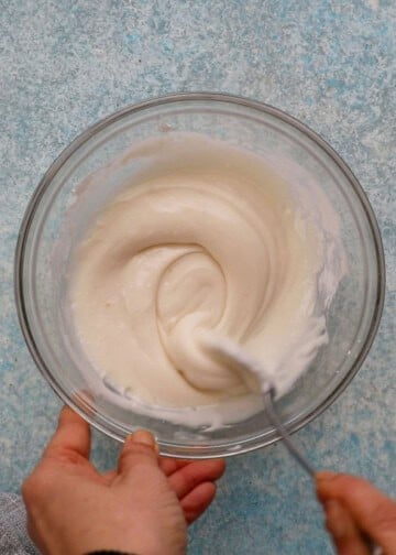 a hand mixing white sugar icing in a small glass bowl using a spoon. 