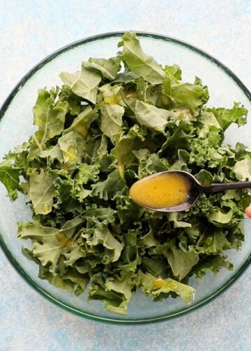 a hand drizzling yellow salad dressing over chopped green kale in a glass bowl.