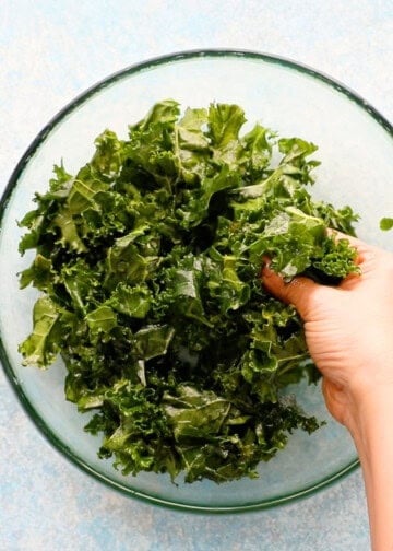 a hand drizzling yellow salad dressing over chopped green kale in a glass bowl.