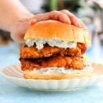 a hand pressing one tandoori chicken sandwich on a white plate.