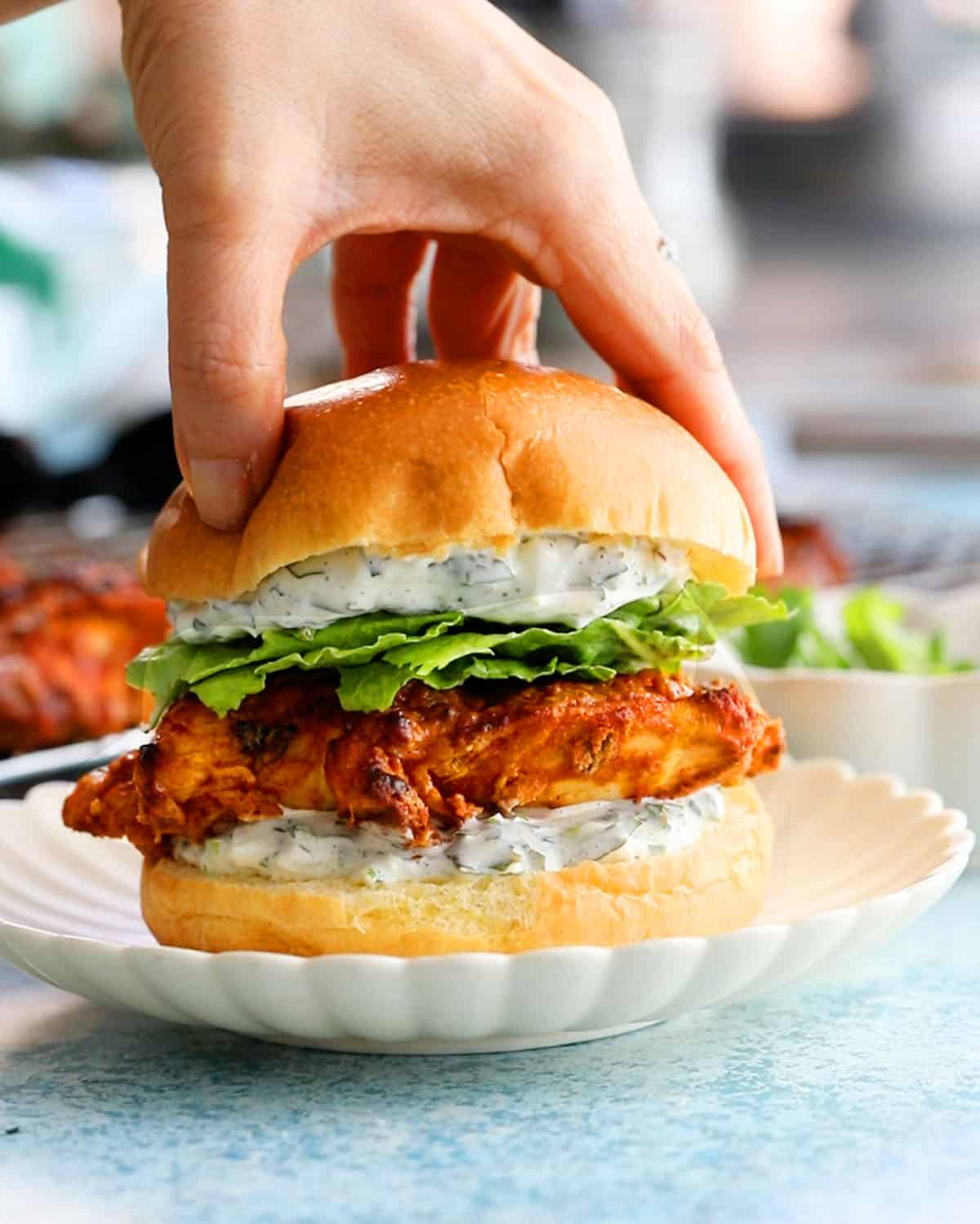 a hand pressing on tandoori chicken sandwich on a white plate.