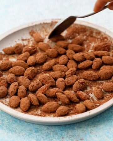 a hand mixing almonds in cocoa powder in a plate using a black spoon.