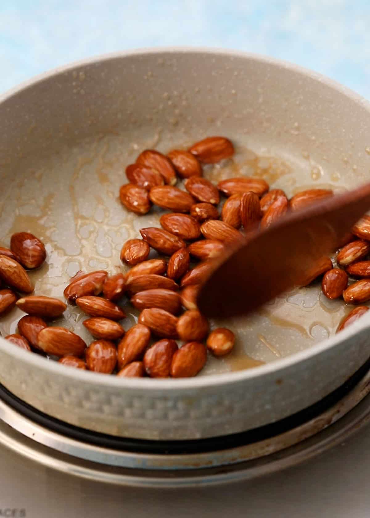 a hand stirring almonds in a white skillet using a brown spoon.