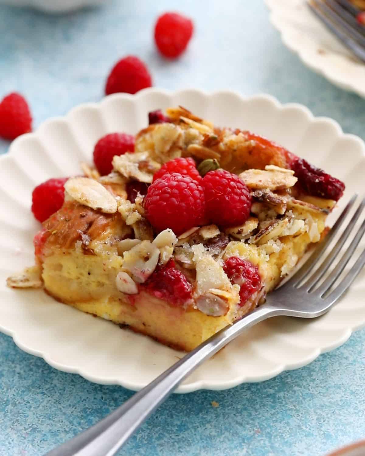 one slice of baking raspberry french toast casserole in a fluted white plate along with a fork.