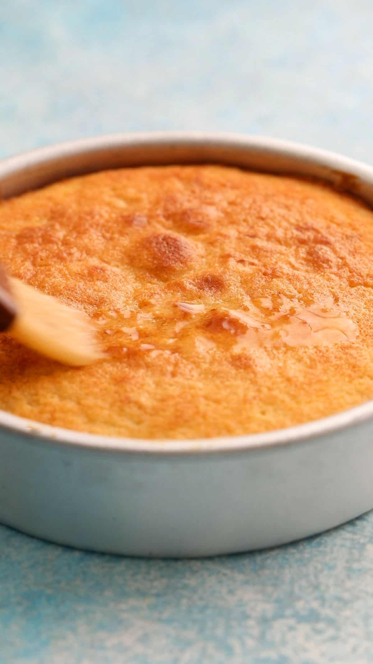 a hand brushing lemon syrup on top of a brown cake in a cake pan.