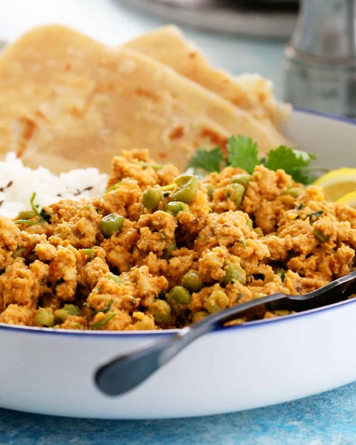 brown chicken keema with green peas in a white bowl along with a black spoon.