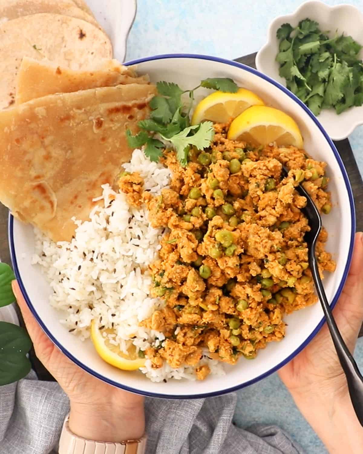 two hands holding one white bowl with brown chicken keema and green peas.