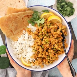 two hands holding one white bowl with brown chicken keema and green peas.