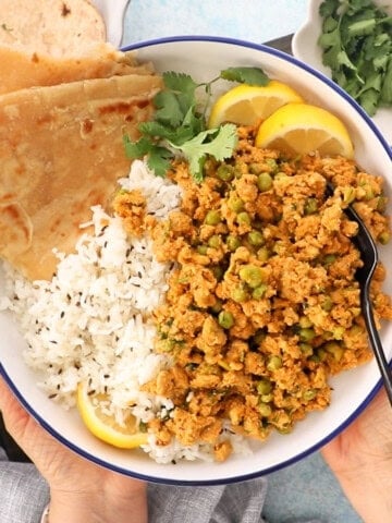 two hands holding one white bowl with brown chicken keema and green peas.