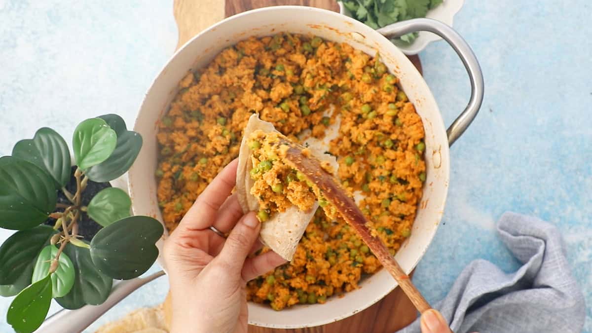 two hands scooping up soem chicken keema into a piece of brown flat bread.