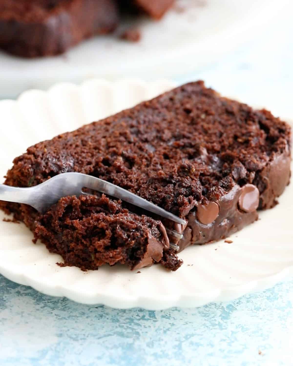 a fork cutting into a slice of brown chocolate zucchini bread in a white plate.