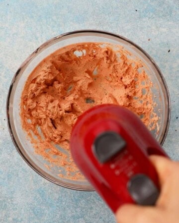 a red beater beating pink cream cheese in a glass bowl.